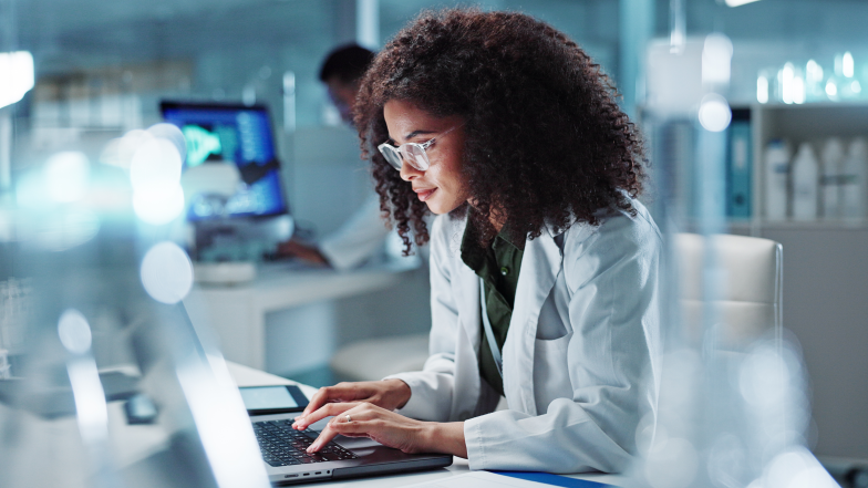 woman in lab coat reviews information on a laptop surrounded by laboratory equipment